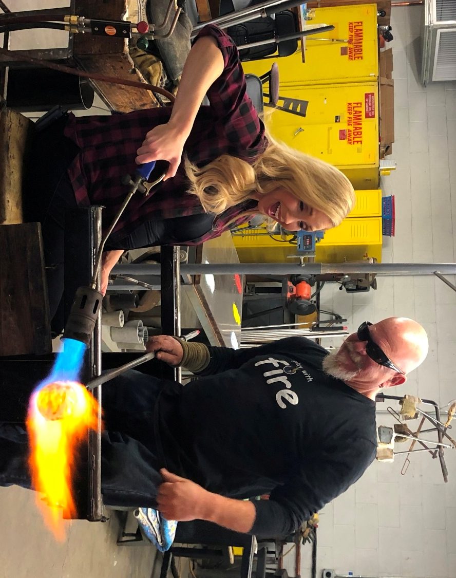 glass blowing class woman on the bench with torch, instructor looking on